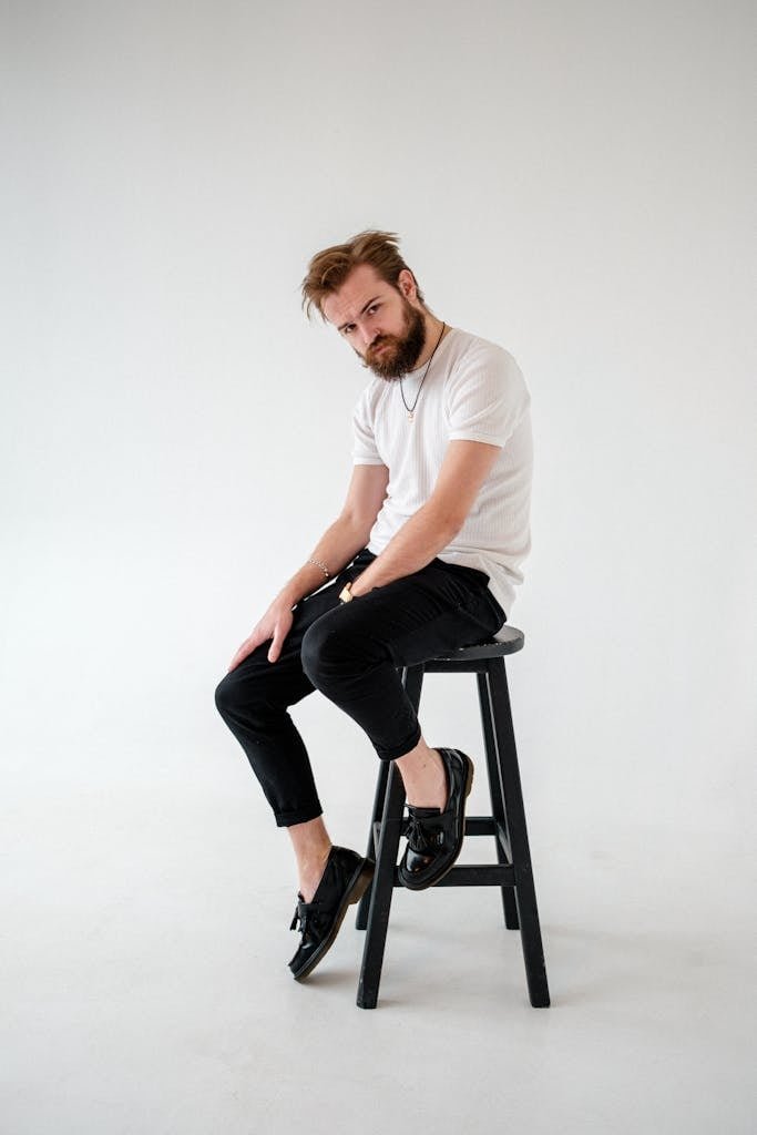Cool young male with beard in t shirt and leather shoes looking at camera while sitting on stool on light background