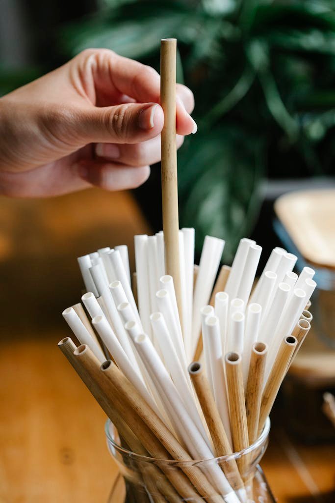 Crop anonymous person picking eco friendly bamboo straw from glass jar placed on wooden table in light kitchen