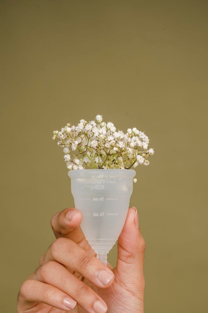 Crop unrecognizable female demonstrating reusable menstrual cup with small fresh white flowers against beige background