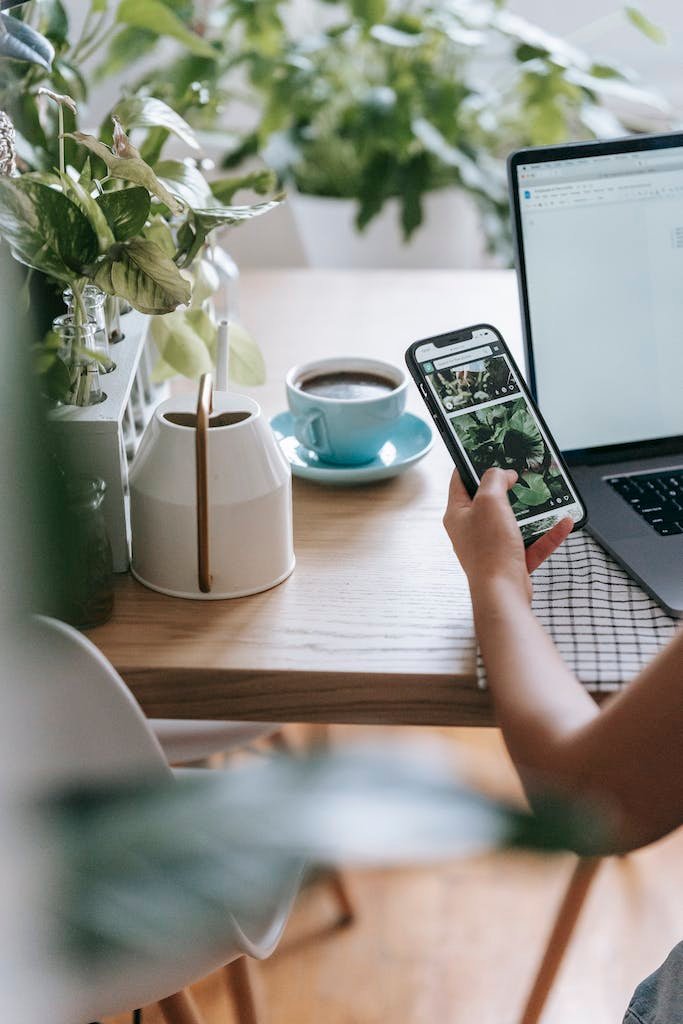 Crop unrecognizable person working on modern netbook and using mobile phone while sitting at desk with potted plants and cup of coffee