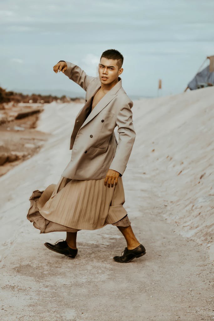 Side view of stylish ethnic male in jacket and pleated skirt walking on dry land while looking at camera